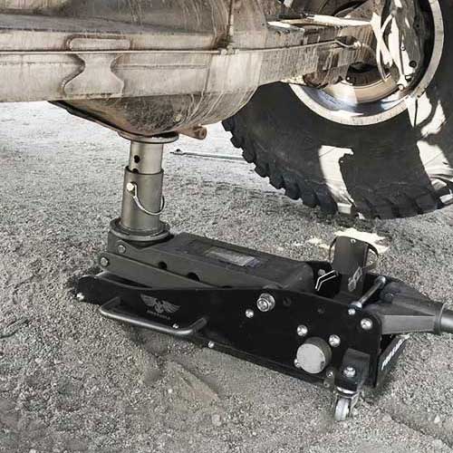 Black 2 Ton off-road jack supporting a truck's undercarriage on a gravel surface.